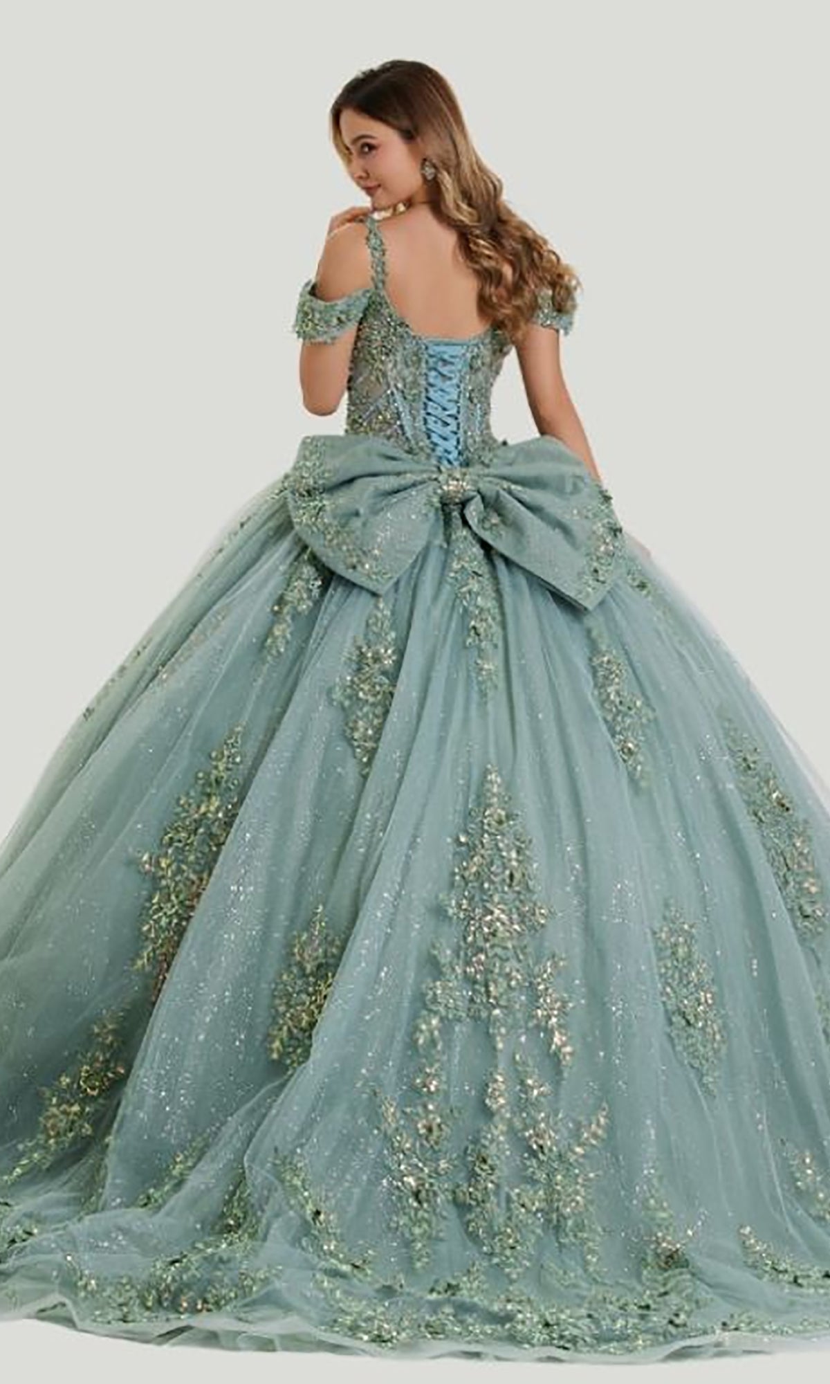 A young woman stands facing away and looks over her shoulder, wearing the Quinceanera Dress 26403 by Stella Couture—a stunning, light blue gown with an embellished corset bodice, a large bow accent, intricate floral details, and a glittering tulle skirt.